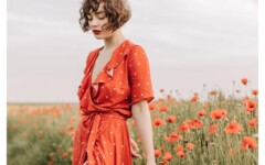 Žena s kovrčavom smeđom kosom u crvenoj haljini s bijelim točkicama stoji u polju makova
