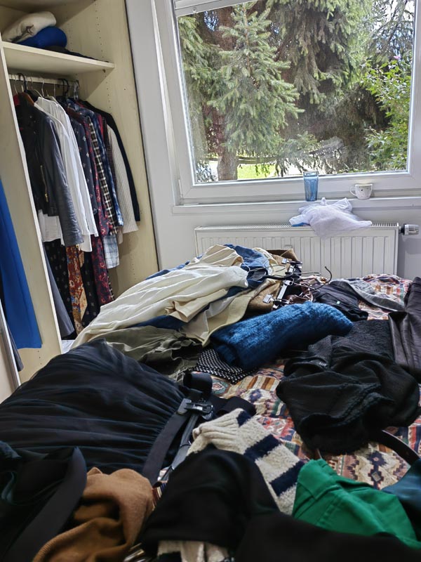 razbacana-odjeca-na-krevetu-ciscenje-ormara Gomila razbacane i šarene odjeće na krevetu pokraj otvorenog ugradbenog ormara s odjećom na vješalicama i velikog prozora s pogledom na drveće.