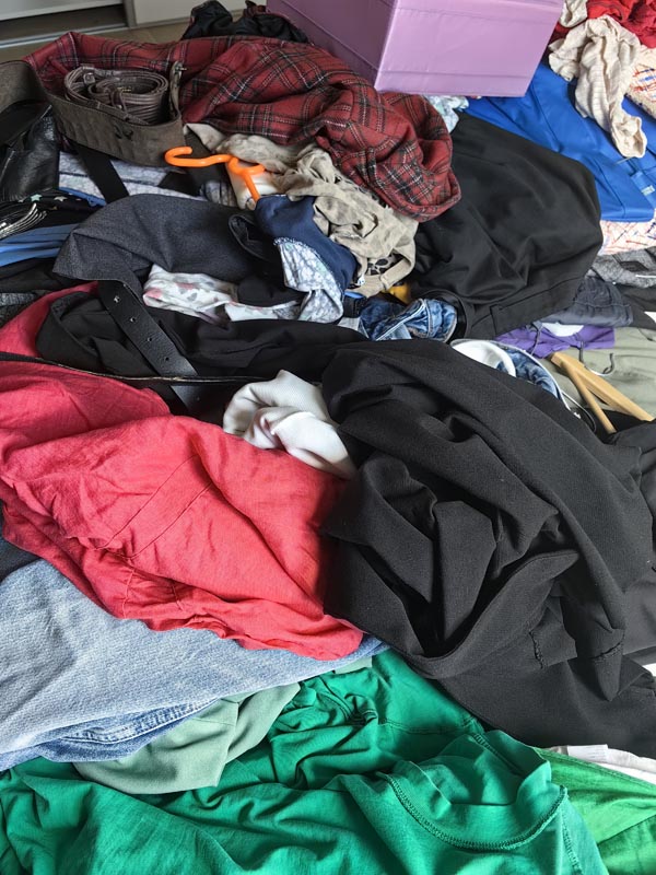 velika-gomila-nerazvrstane-odjece-na-podu Krupni kadar velike gomile razbacane ženske odjeće raznih boja, uključujući crne hlače, crvenu košulju, zelenu majicu i kariranu jaknu, spremne za sortiranje.