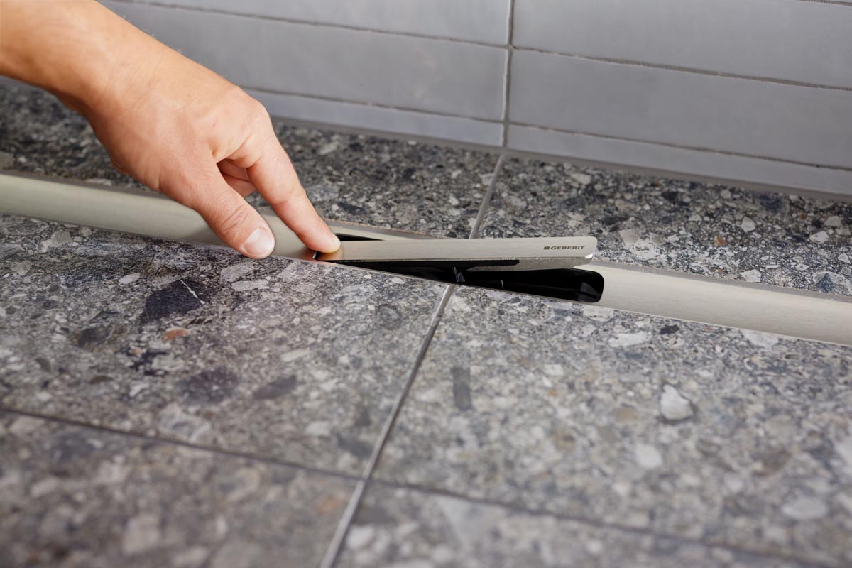 Ruka podiže metalni poklopac linijske tuš kanalice Geberit CleanLine na kamenom podu walk-in tuša.