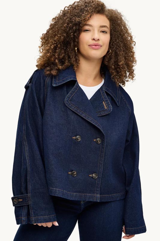 kratka-traper-jakna-dvoredno-kopcanje-oversized Žena s voluminoznom kovrčavom kosom nosi tamnoplavu oversized kratku traper jaknu s dvorednim kopčanjem, širokim ovratnikom i remenčićima na manžetama preko bijele majice.