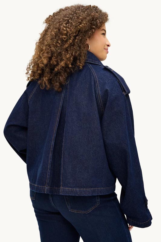 kratka-traper-jakna-oversized-prikaz-s-leda Prikaz s leđa tamnoplave oversized kratke traper jakne s velikim naborom na leđima, spuštenim ramenima i epoletama, koju nosi žena s kovrčavom kosom.
