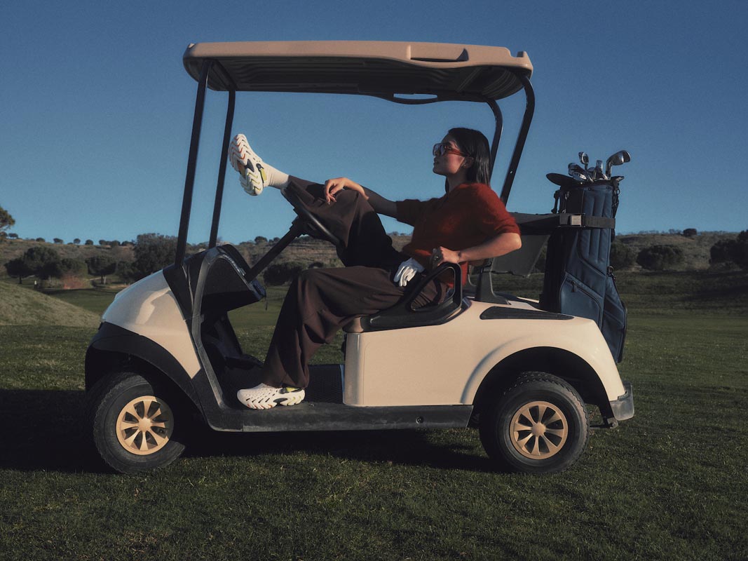 Ženske natikače Crocs Echo II u bijeloj boji na nogama modela koji sjedi u vozilu za golf na travnatom terenu.
