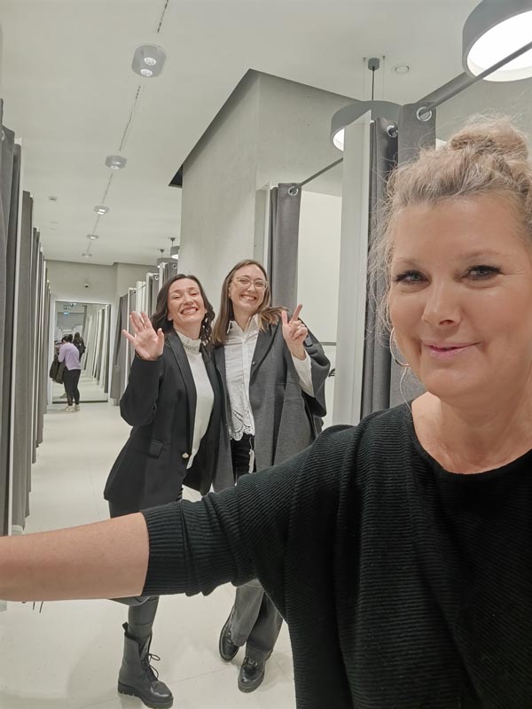 Tri nasmijane žene snimaju selfie u hodniku s kabinama za presvlačenje u trgovini odjećom, dok dvije u pozadini veselo mašu i poziraju.