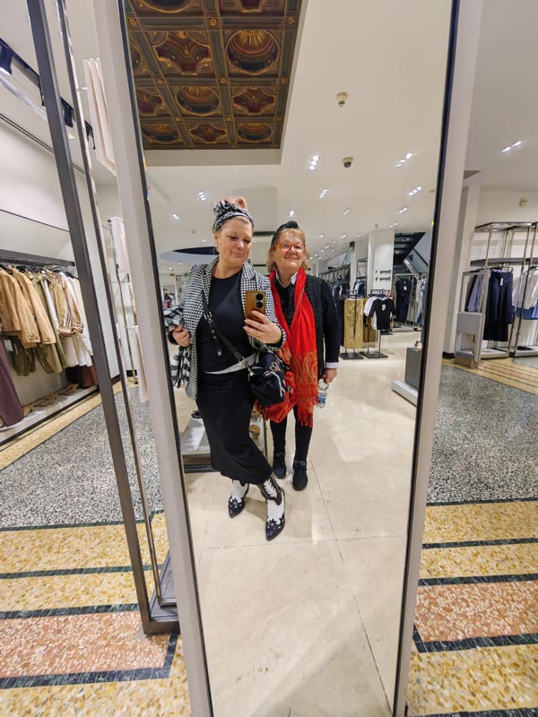 Zajednički selfie dviju žena u velikom ogledalu luksuzne trgovine odjećom; jedna nosi pepito sako, crnu dugu suknju i kaubojske čizme, a druga crnu jaknu s upečatljivim crvenim šalom.