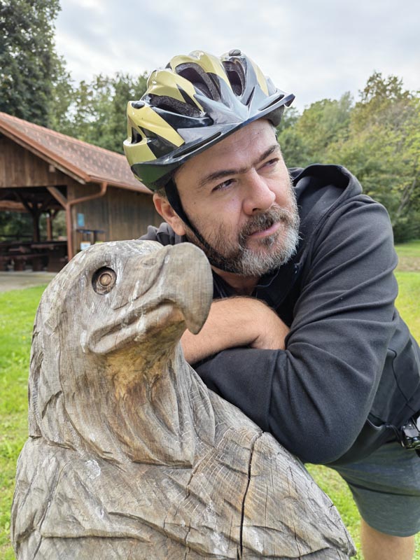 Nikola Zoko s biciklističkom kacigom naslonjen na drvenu skulpturu orla na otvorenom