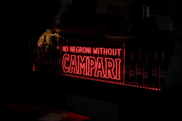 Crveni neonski natpis s porukom No Negroni Without Campari svijetli u mračnom baru pokraj izloženih boca pića na šanku.