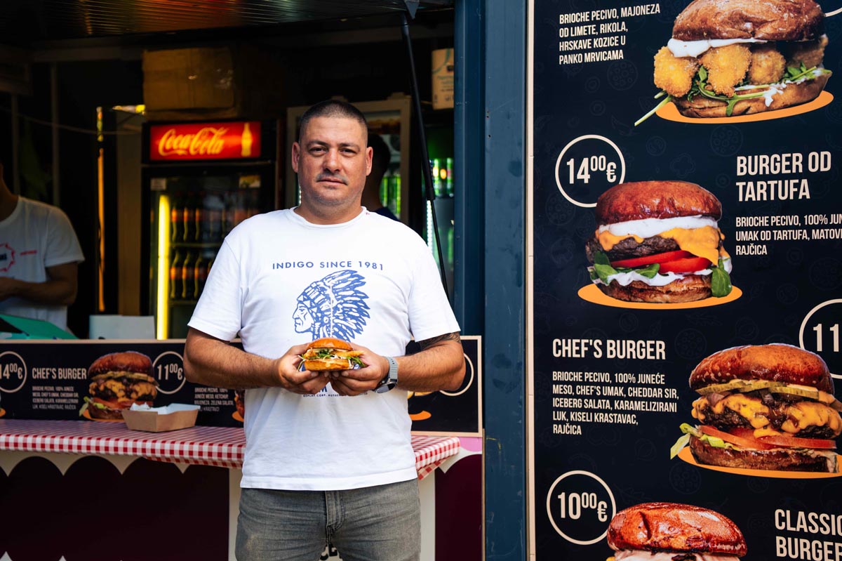 Tomislav Strikoman drži burger ispred festivalske kućice s cjenikom i ponudom burgera