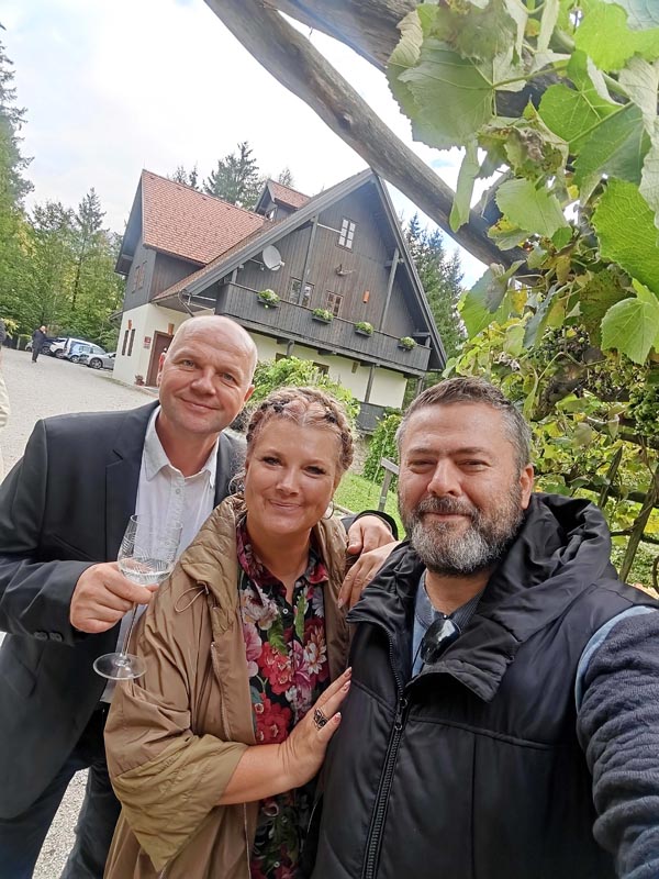 Alma, Janko i Nikola snimaju selfie ispred tradicionalne drvene kuće u prirodi, muškarac s lijeve strane drži čašu vina.