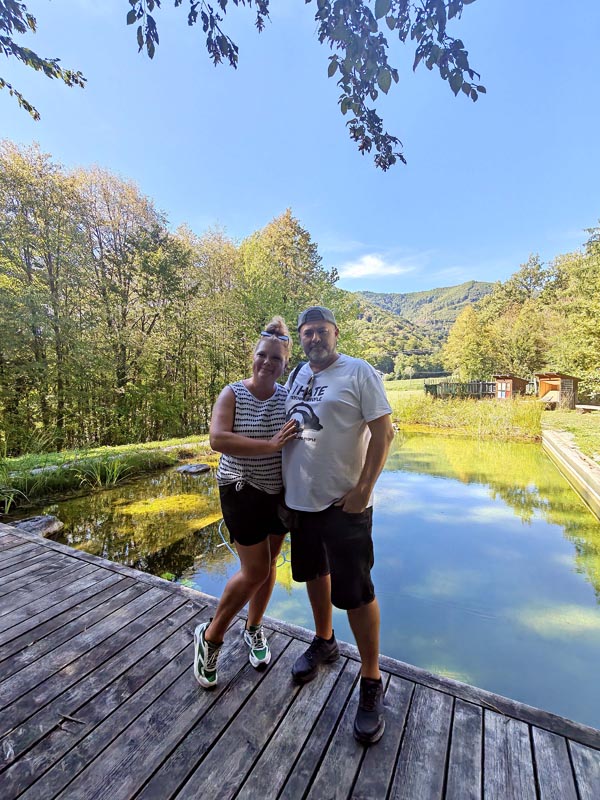 Alma i Nikola stoje zagrljeni na drvenom molu pokraj bistrog jezera, okruženi zelenom šumom i brdima u pozadini pod plavim nebom.