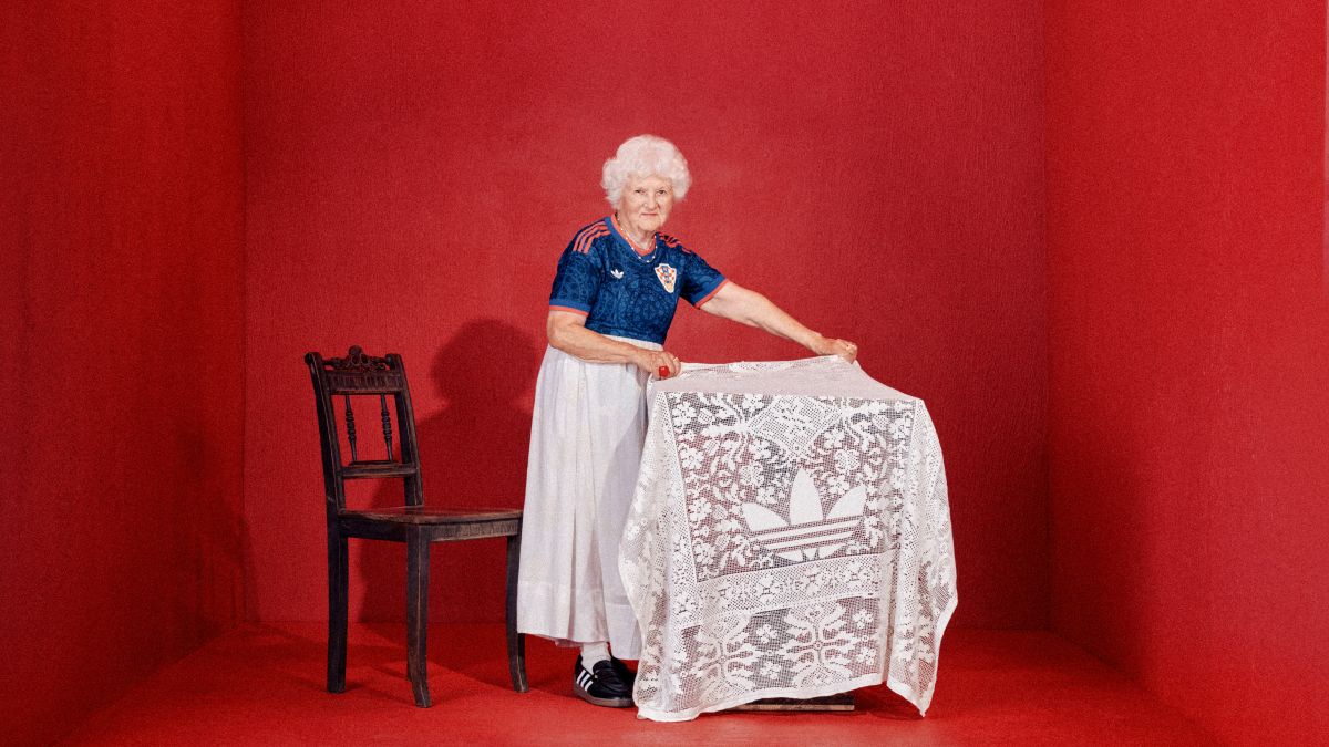 Starija žena u plavom hrvatskom dresu širi tradicionalni stolnjak s motivom čipke i logom brenda Adidas
