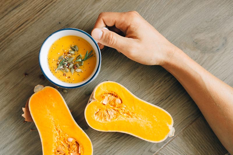 Gusta krem juha od bundeve u šalici ukrašena bučinim sjemenkama pored prerezane butternut tikve.