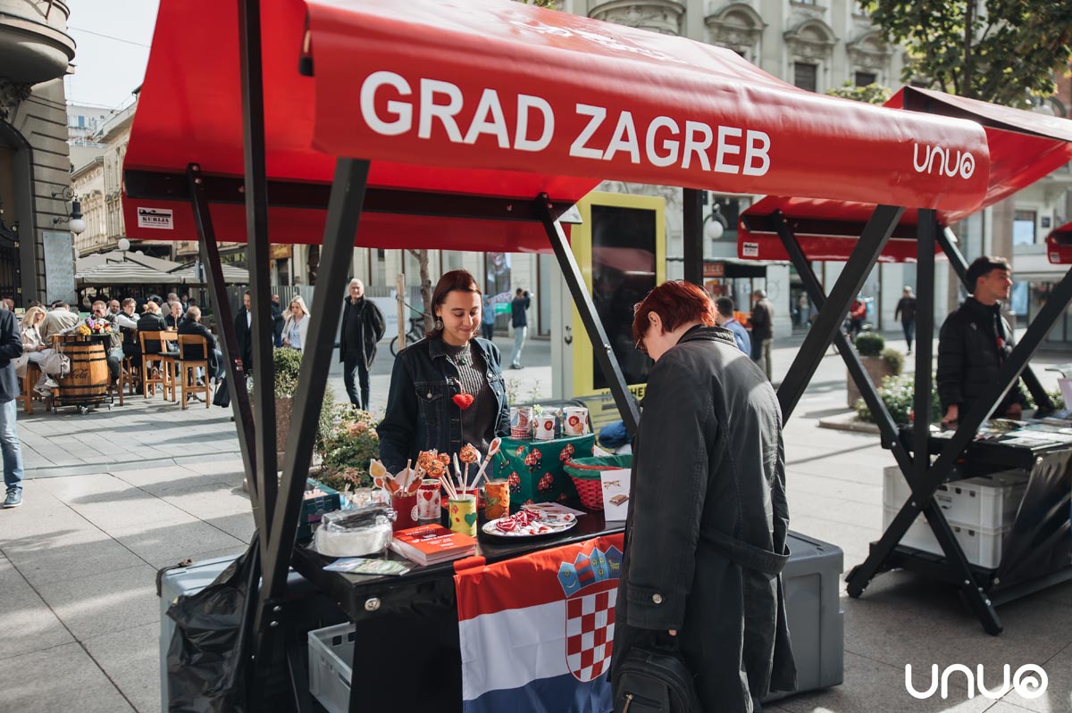 Prodajni štand udruge UNUO pod crvenom tendom s natpisom Grad Zagreb na gradskom trgu
