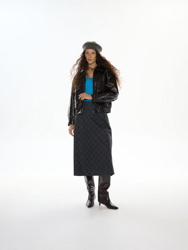 Žena s beretkom u Reserved lakiranoj jakni, kariranoj suknji i kožnim čizmama na bijeloj pozadini