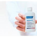 Ruka drži medicinski šampon protiv peruti Ducray Kelual DS Intensive ispred bijele pozadine