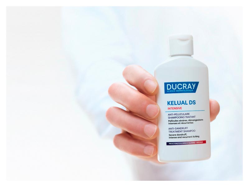 Ruka drži medicinski šampon protiv peruti Ducray Kelual DS Intensive ispred bijele pozadine