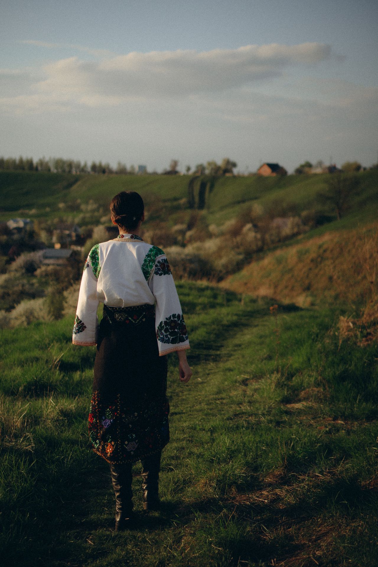 Pogled s leđa na ženu u tradicionalnoj narodnoj nošnji s crnom suknjom i vezenom bluzom kako hoda zelenim brežuljkom
