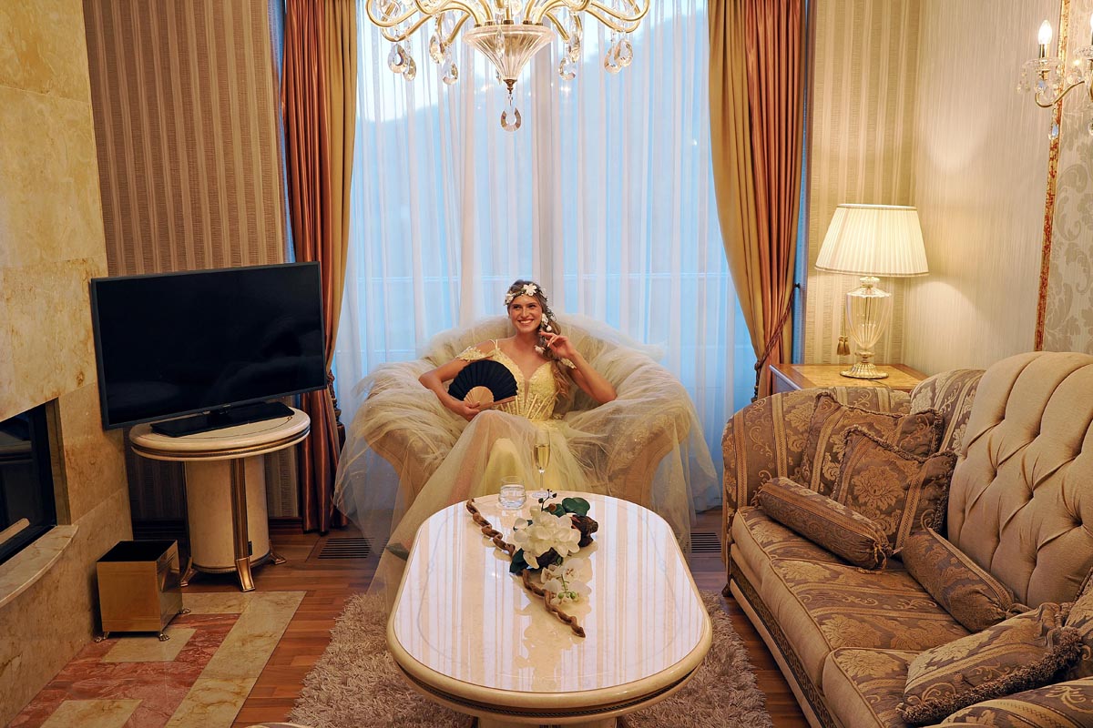 Nasmijana žena u svečanoj žutoj haljini s lepezom sjedi u raskošnoj fotelji hotelskog apartmana