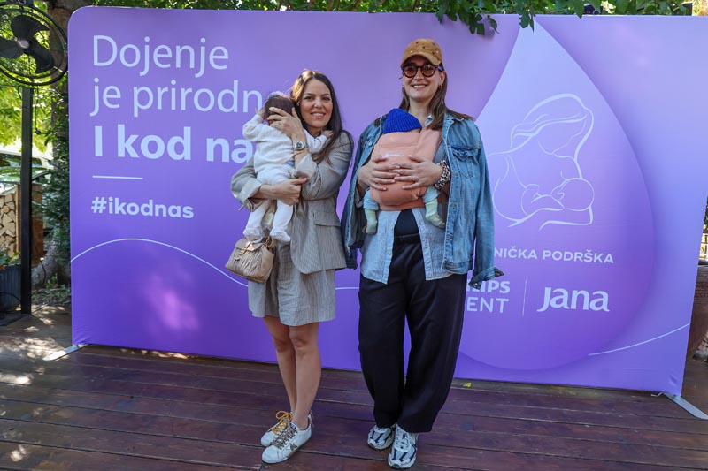 Dvije majke s bebama u naručju i nosiljci ispred panela Dojenje je prirodno I kod nas