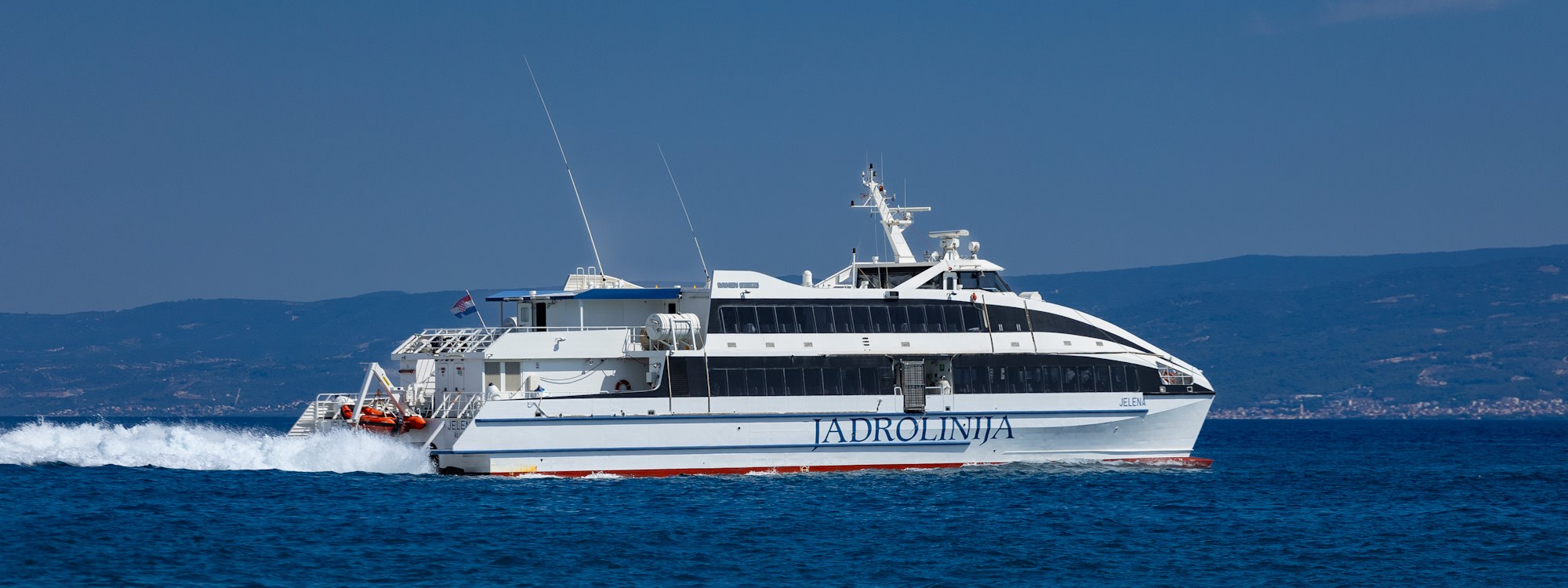 Bijeli brzi katamaran Jelen u vlasništvu tvrtke Jadrolinija plovi plavim Jadranskim morem i ostavlja bijeli pjenasti trag, dok se u pozadini pod vedrim nebom nazire obala s brežuljcima.