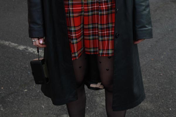 Modni street style outfit s dugim crnim kožnim kaputom, crvenom kariranom suknjom, crnim najlonkama sa srcima i vintage torbicom.