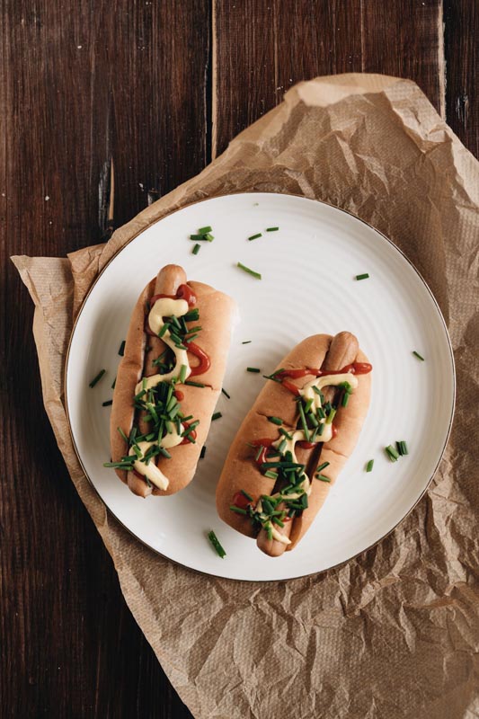 Dva hot doga sa senfom, kečapom i vlascem na bijelom tanjuru i smeđem papiru