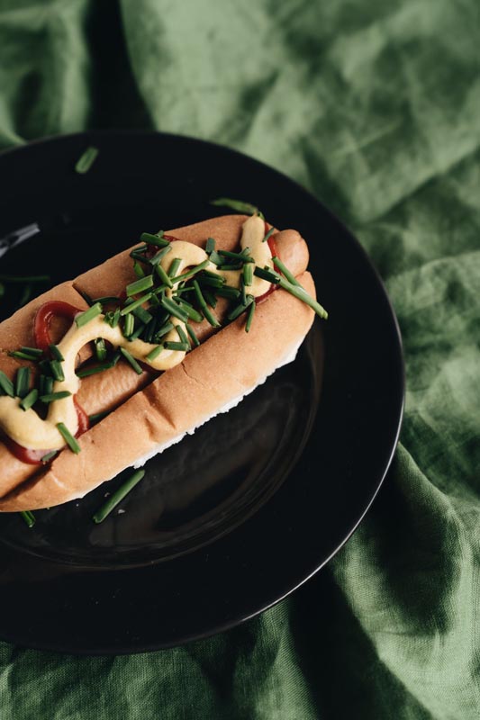 Hot dog sa senfom i narezanim vlascem na crnom tanjuru pokraj zelene tkanine