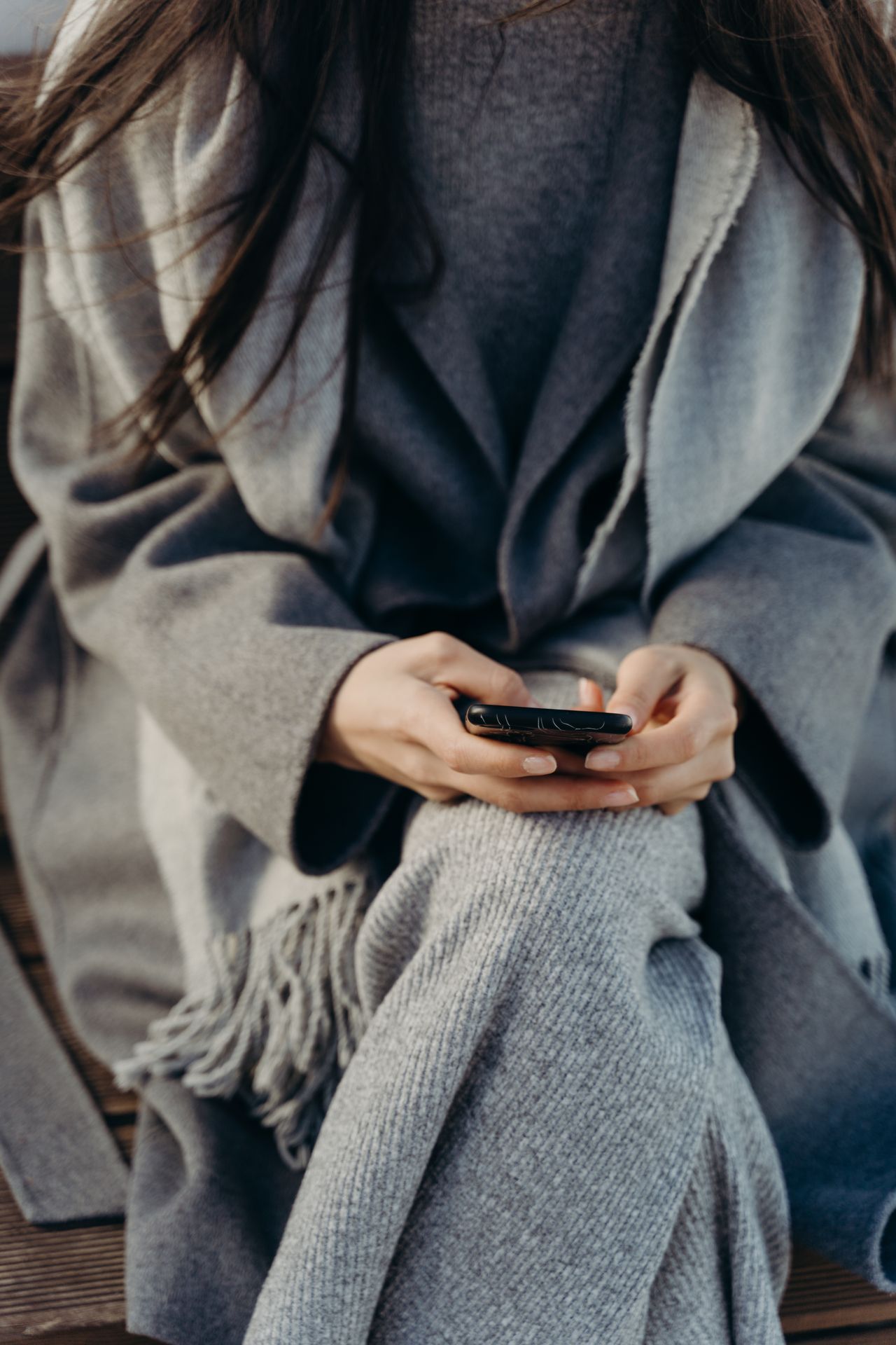 Sivi pleteni šal s resama i topli kaput u rukama djevojke koja drži mobitel