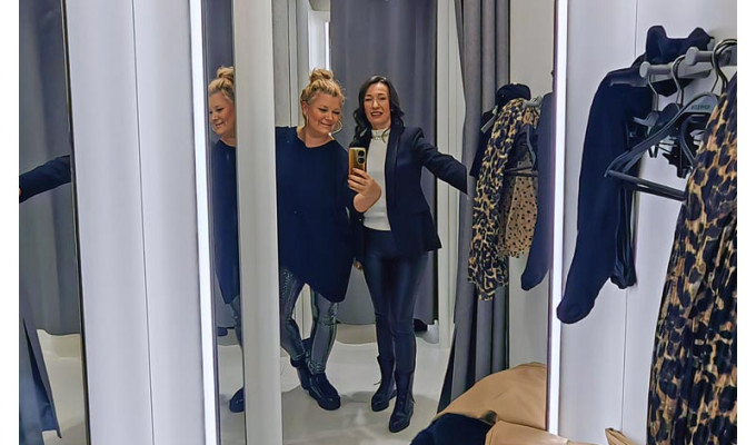 Dvije nasmijane prijateljice snimaju zajednički selfie u ogledalu kabine za presvlačenje u trgovini odjećom, dok se u pozadini vide zavjese i odjeća na vješalicama.