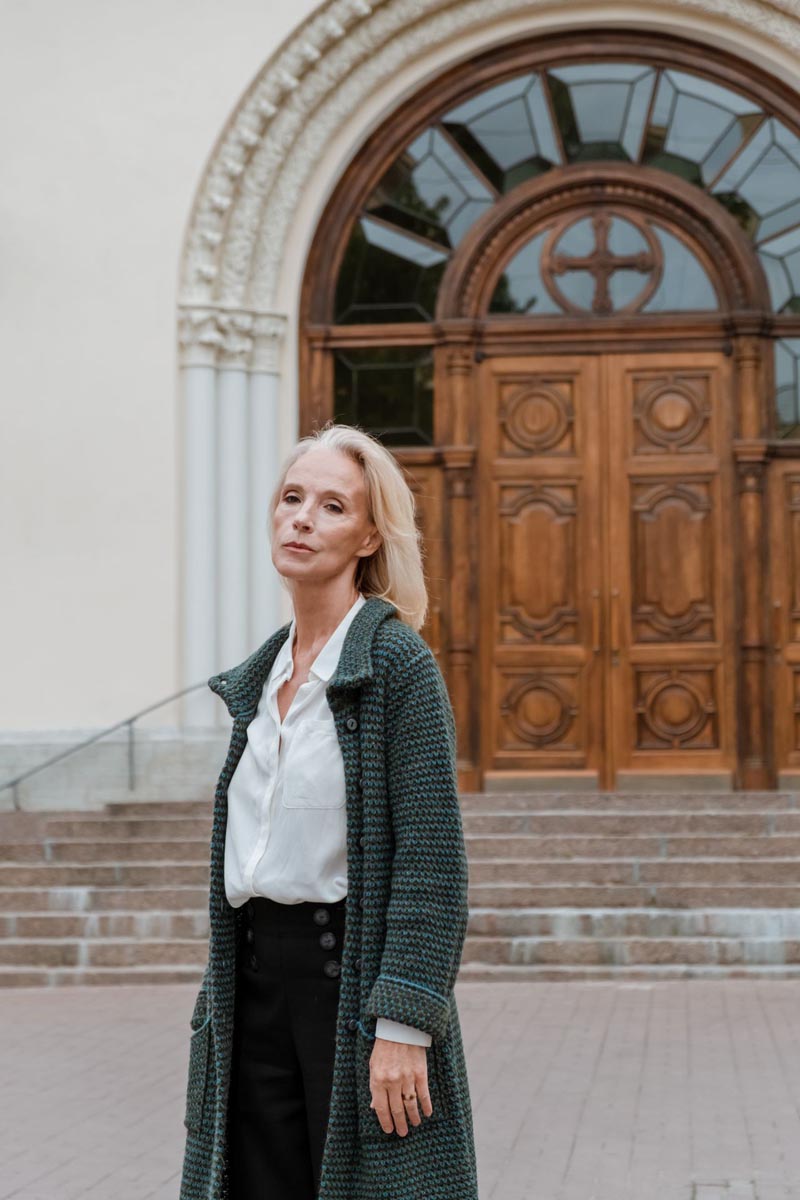 Starija žena u dugom zelenom pletenom kaputu ispred velike drvene crkvene kapije