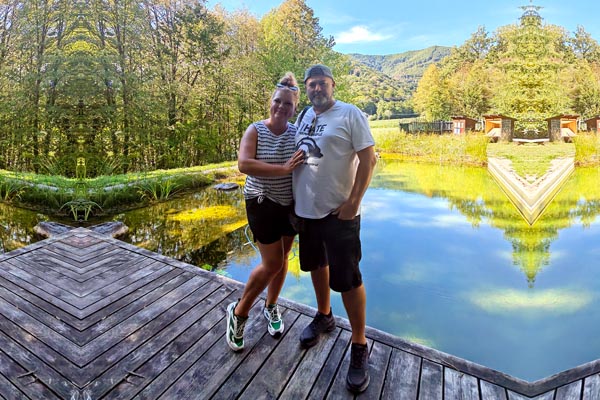 Alma i Nikola Zoko poziraju na drvenom molu pokraj bistrog prirodnog jezera okruženog zelenom šumom i brdima pod plavim nebom.