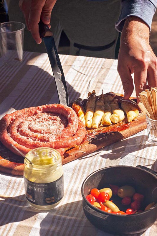Domaća kobasica smotana u spiralu i zapečeni sir na drvenoj dasci uz staklenku umaka i masline na stolu.