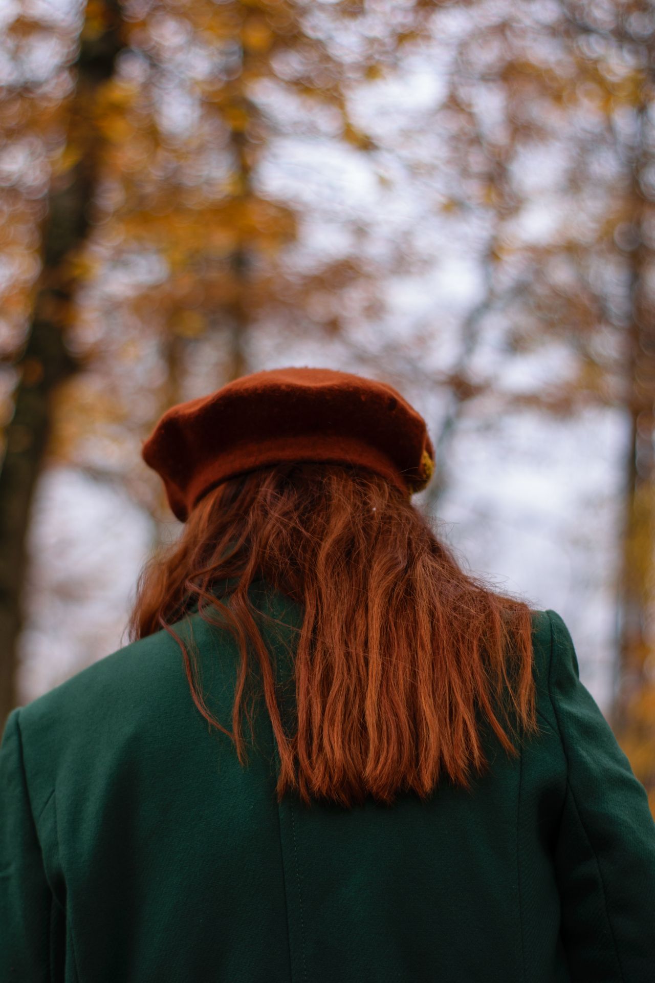 Žena s beretkom i u zelenom kaputu šeta kroz jesenji park okrenuta leđima
