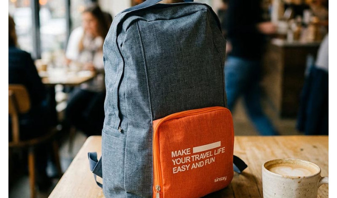 Sinsay sklopivi putni ruksak u sivo-narančastoj boji s natpisom "Make your travel life easy and fun" postavljen na drveni stol u kafiću pokraj šalice kave.