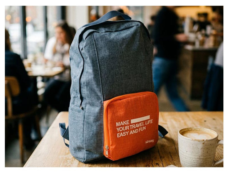 Sinsay sklopivi putni ruksak u sivo-narančastoj boji s natpisom "Make your travel life easy and fun" postavljen na drveni stol u kafiću pokraj šalice kave.