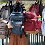 Četiri ruksaka marke JanSport u različitim bojama vise obješeni na metalnoj konstrukciji na otvorenom.