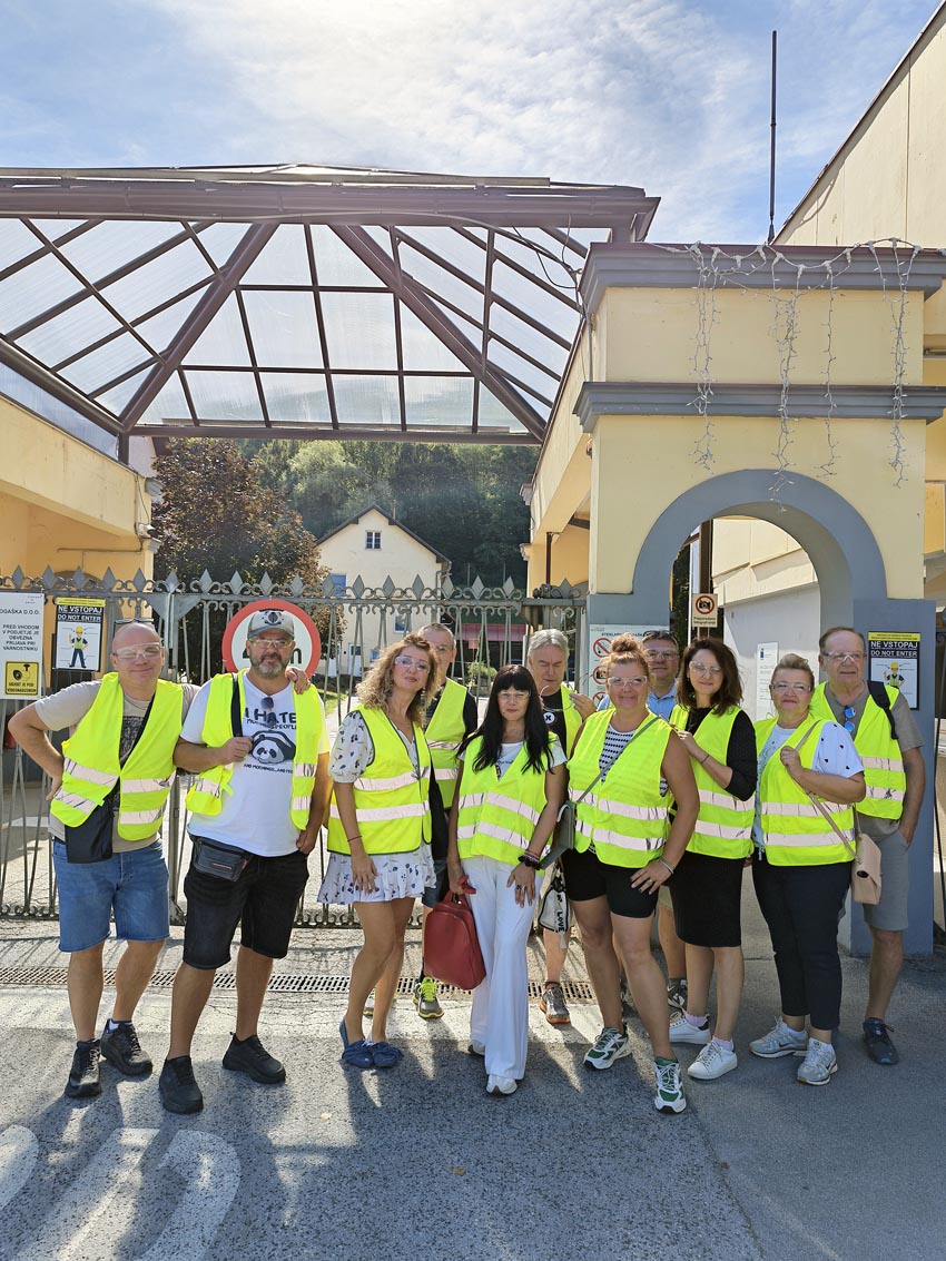 Grupa turista u žutim prslucima pozira ispred ulaza u tvornicu Staklarna Rogaška tijekom organiziranog izleta u Sloveniji.