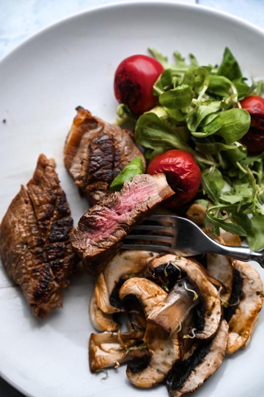 steak_u_restoranu_1 Komad medium pečenog odreska na vilici s prilogom od pirjanih gljiva i svježe salate na bijelom tanjuru