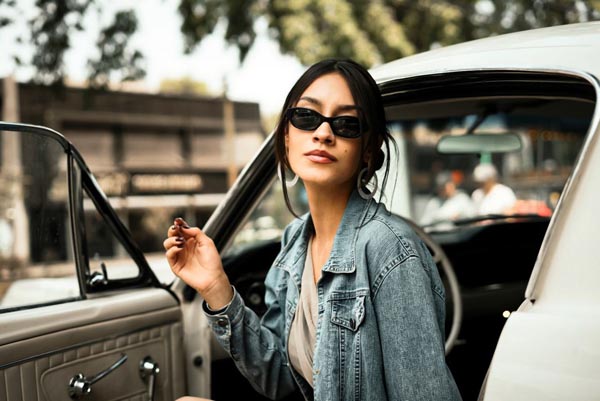 Djevojka s crnim sunčanim naočalama i u traper jakni sjedi na vozačkom mjestu starog automobila