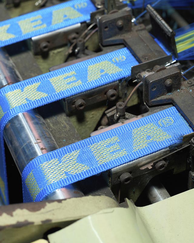 Plave tekstilne trake s prepoznatljivim žutim IKEA logotipom prolaze kroz tvornički stroj za tkanje