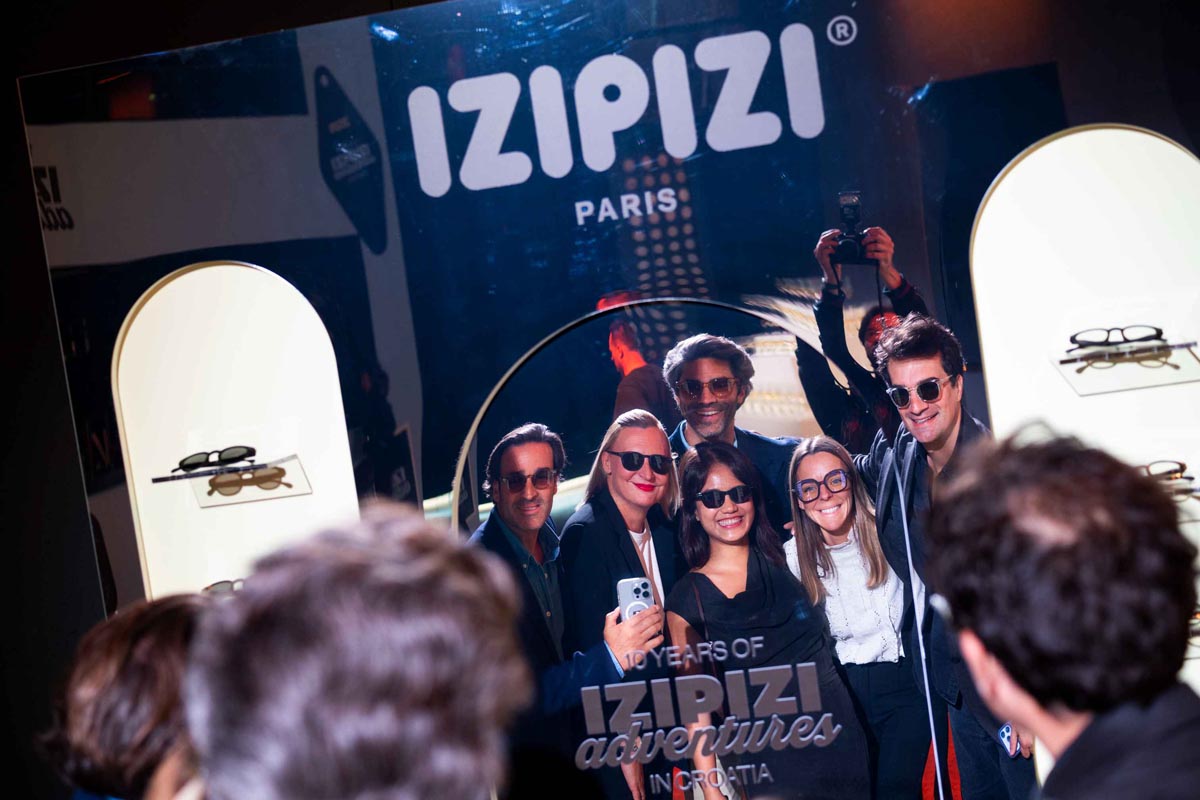 Grupa nasmijanih ljudi sa sunčanim naočalama pozira ispred IZIPIZI logotipa na proslavi