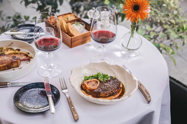 Postavljen stol u restoranu Le Bistro Esplanade s čašama crnog vina, mesnim jelom i narančastim gerberom