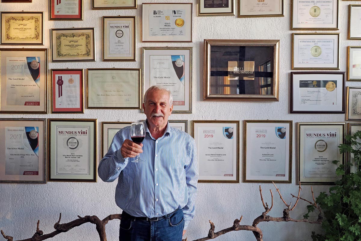 Vinar Nikola Zikatanov s čašom crnog vina ispred zida s brojnim priznanjima i nagradama vinarije Villa Melnik