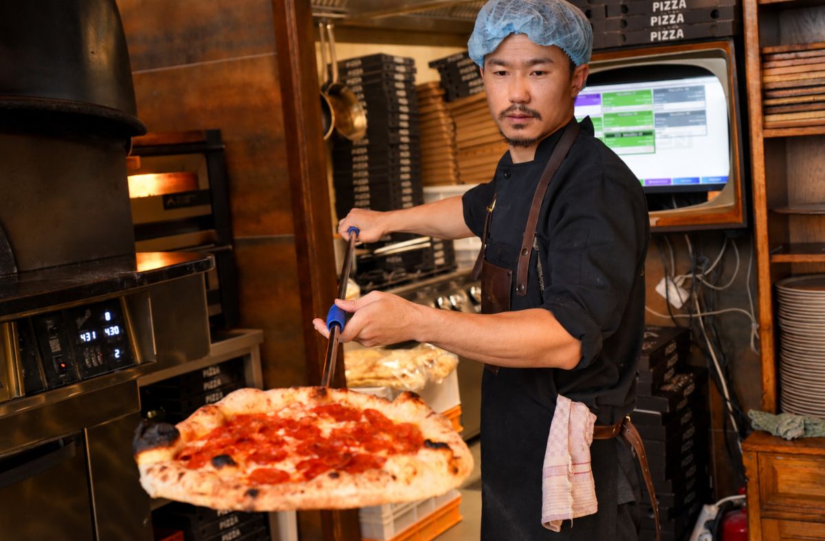 Pizzaiolo s kapom za kosu vadi svježe pečenu pizzu s kulenom iz krušne peći u pizzeriji Mrs. Fogg