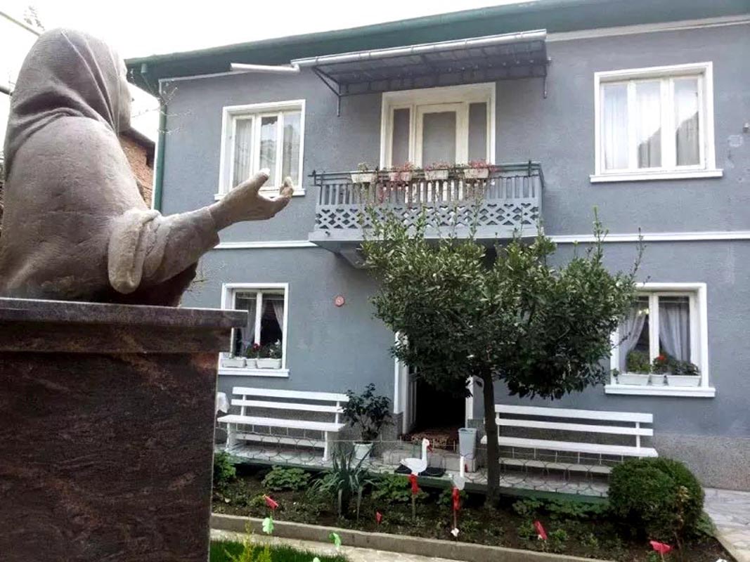 Brončani spomenik ispred ulaza u dvokatnu kuću-muzej Babe Vange s bijelim klupama u Petriču