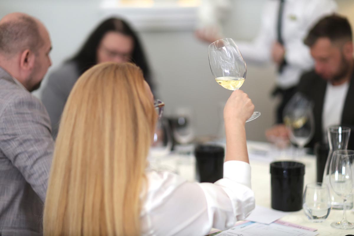 Plavokosa žena s leđa podiže čašu bijelog vina prema svjetlu tijekom profesionalnog ocjenjivanja