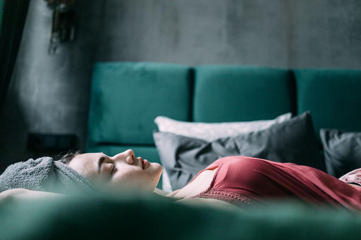 Žena s ručnikom na glavi spava na krevetu s tamnozelenim uzglavljem u crvenoj spavaćici