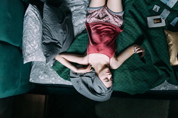 Žena sa sivim ručnikom na glavi leži na krevetu u crvenoj satenskoj pidžami i drži mobitel