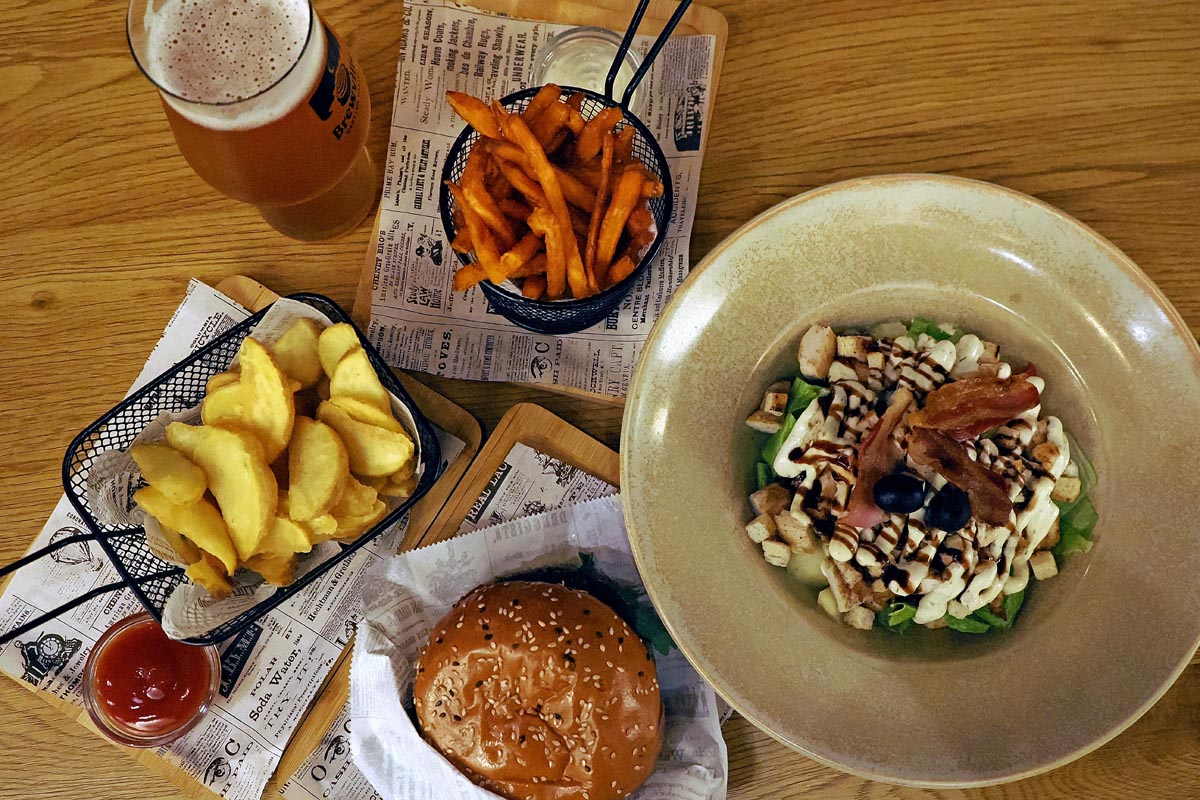 Sočni burger sa sezamom, salata s piletinom i slaninom, krumpirići i pivo na drvenom stolu u Brewville pubu u Metkoviću