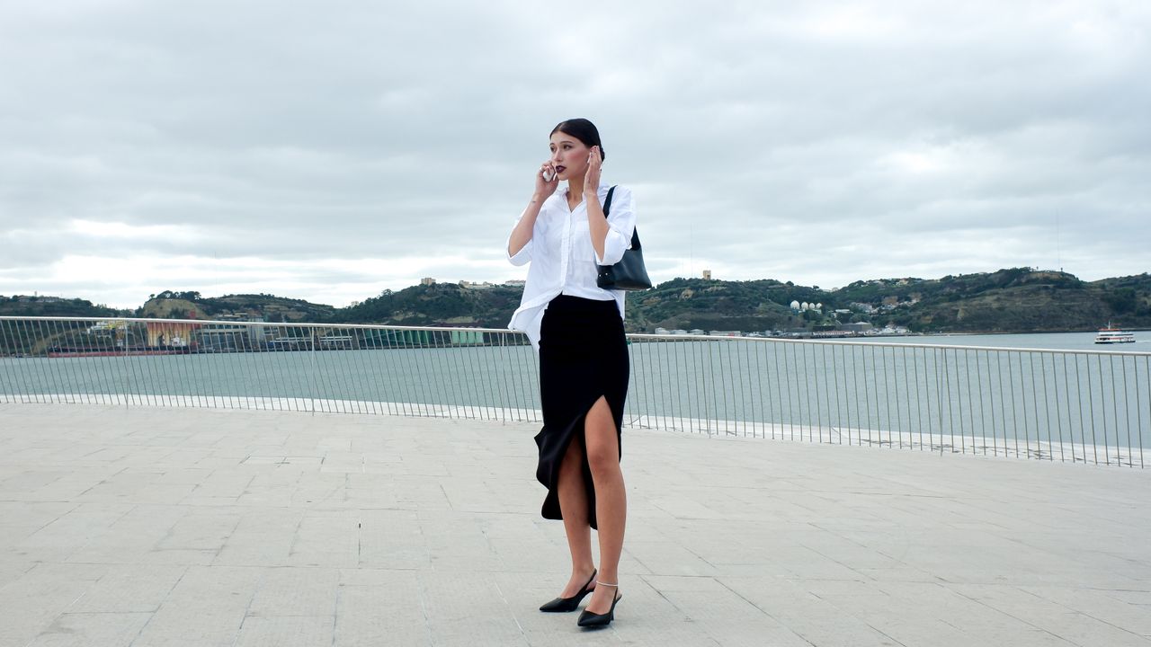 Mlada žena razgovara na mobitel s bežičnim slušalicama, kombinirajući bijelu košulju i crnu suknju za moderan poslovni look
